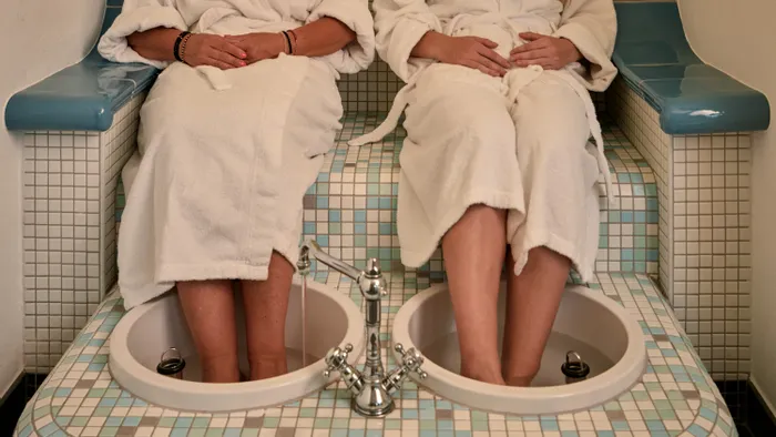 Zwei Frauen in weißen Bademänteln genießen ein entspannendes Fußbad in gefliestem Spa-Bereich